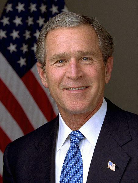 Photo of George W. Bush. He is smiling and standing in front of an American flag while wearing a suit.